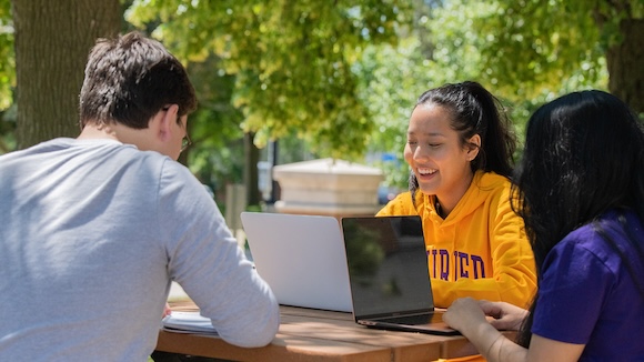 Students sitting at table working on laptops under trees