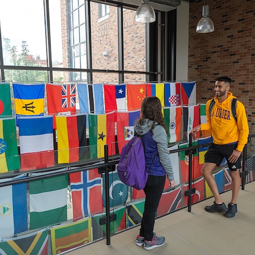 Two students having a conversation on a balcony with strings of flags from the world hanging in front of a large window