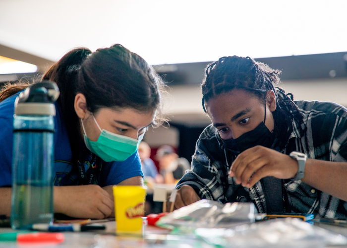 Two masked females sitting a table working on a project together.